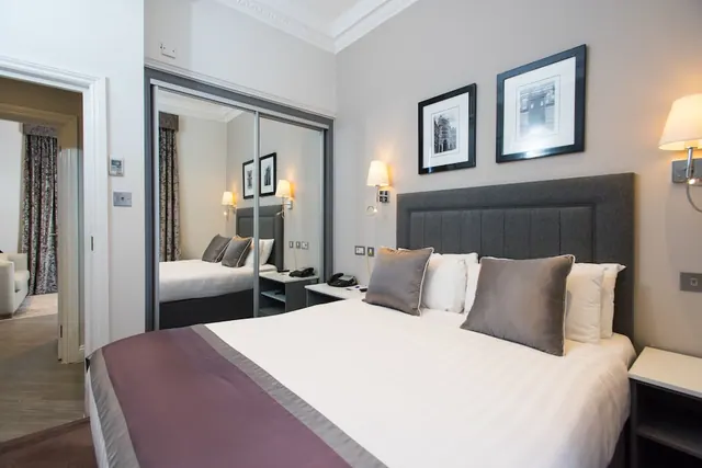 A made-up double bed with a grey upholstered headboard and cushions sits beside a mirrored sliding wardrobe, with framed prints and wall lamps on the wall.