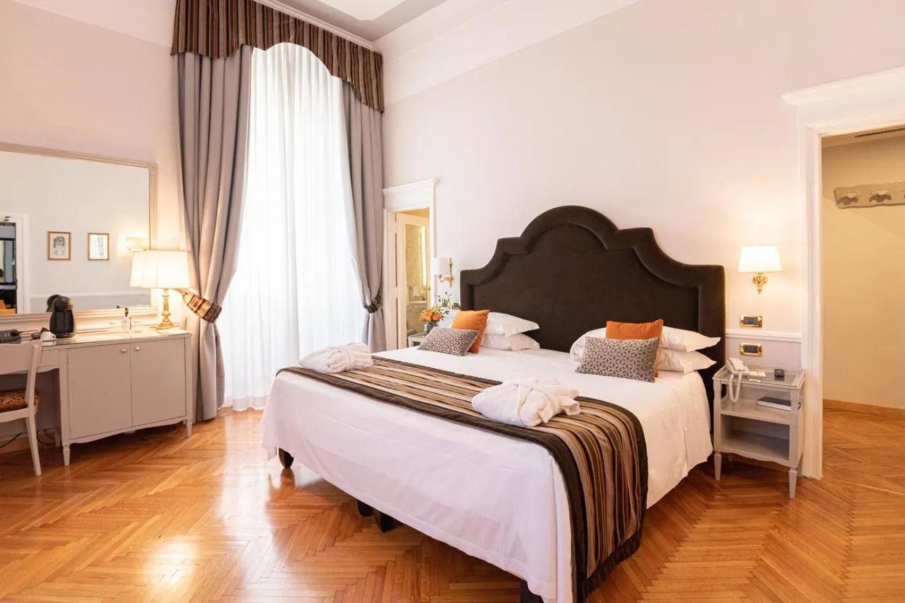 A bedroom with a made king bed with a dark upholstered headboard, a striped runner and folded white robes, beside a dressing table and a tall arched window with sheer curtains.