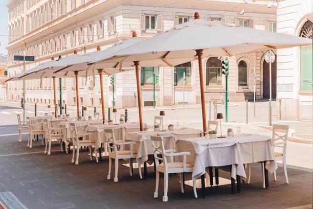 An empty outdoor dining terrace with white umbrellas, white-clothed tables and white chairs lines a street in front of a pale stone building.