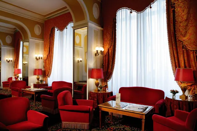 A hotel lounge with red velvet armchairs, a red sofa, wood coffee tables with newspapers, and sheer white curtains over tall windows.