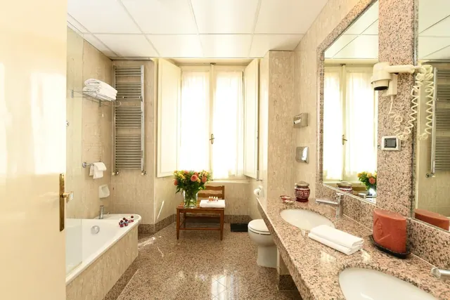 A bathroom with a built-in bathtub, a double vanity with a granite countertop, a toilet, and a window with open shutters and a vase of flowers.