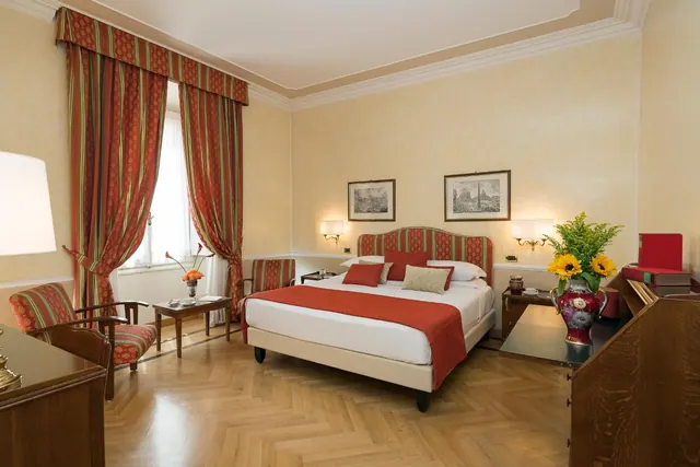 A bedroom with a double bed under red and white bedding, striped red curtains at the window, two armchairs, and a wooden desk holding a vase of sunflowers.