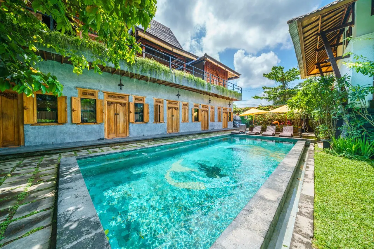 A rectangular turquoise swimming pool sits in front of a two-storey blue building with wooden shutters and doors, with sun loungers and yellow umbrellas at the far end.