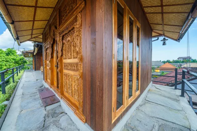 A carved timber villa facade with tall wooden-framed windows, a stone-paved terrace and a thatched overhang, with rooftops and trees beyond.