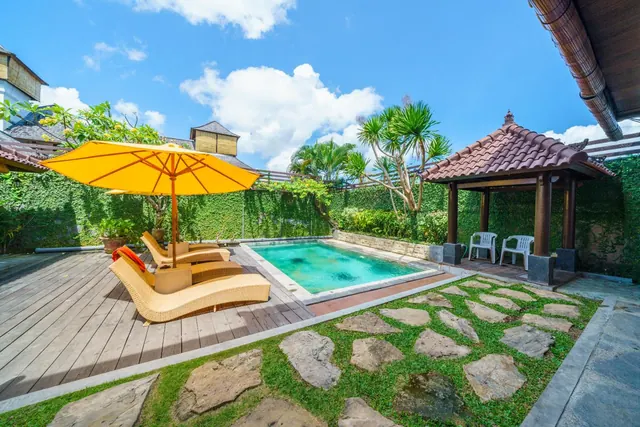 A small private swimming pool sits in a walled garden with a wooden deck, a yellow umbrella, wicker sun loungers and a tiled gazebo.