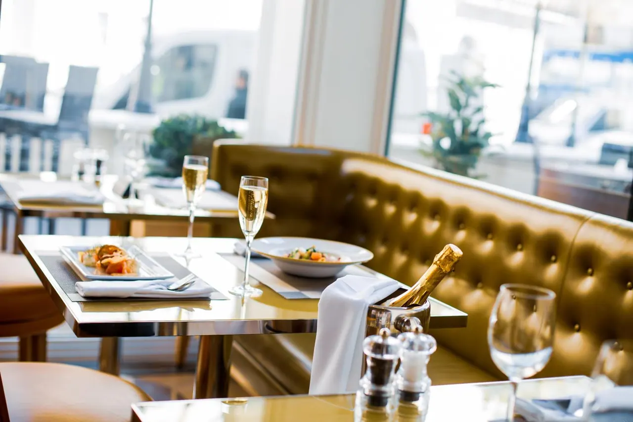 A restaurant table is set with champagne flutes, a champagne bottle in an ice bucket and a plated dish, beside a tufted leather banquette, with a street visible through the window.