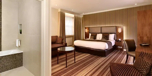 A hotel bedroom with a king bed, a brown sofa and a small round table, with an open bathroom and bathtub on the left.