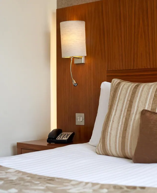 A made bed with striped pillows sits beside a wooden headboard panel, a lit wall lamp, and a black telephone on a bedside table.