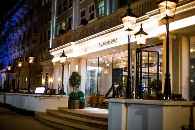 Night exterior of the Blakemore hotel entrance with a white classical facade, lit lamp posts, and potted topiary beside the front steps.