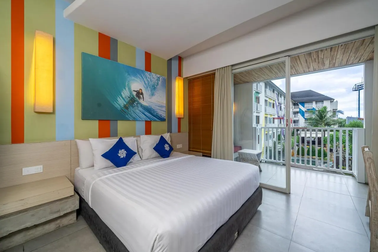 A bedroom with a king bed, a surfing artwork above the headboard, and sliding doors onto a balcony overlooking palm trees and a neighbouring building.
