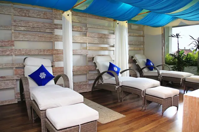Four wicker loungers with white cushions and blue pillows sit under a blue fabric canopy beside a wooden slat wall, with a window showing plants outside.