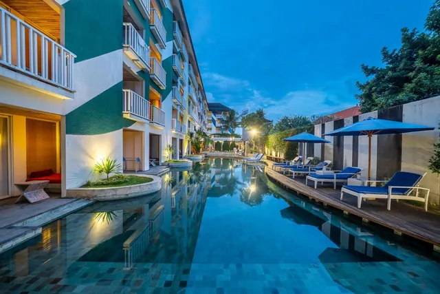 A long swimming pool at dusk with blue sun loungers and umbrellas on a wooden deck beside a multi-storey building with balconies.