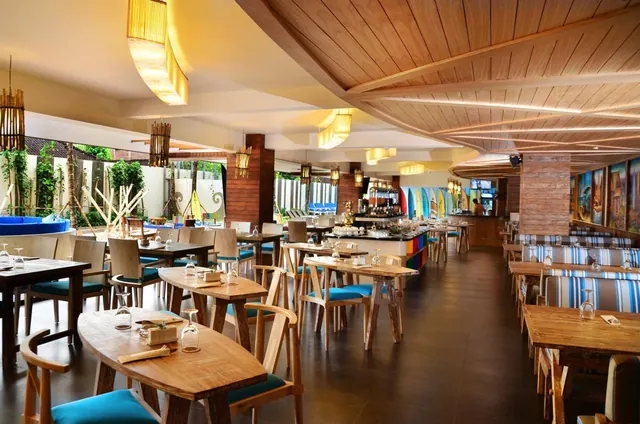 A restaurant dining room with wooden tables, teal chairs, a wood-panelled ceiling and surfboards displayed behind the bar, with a pool area visible through the windows.