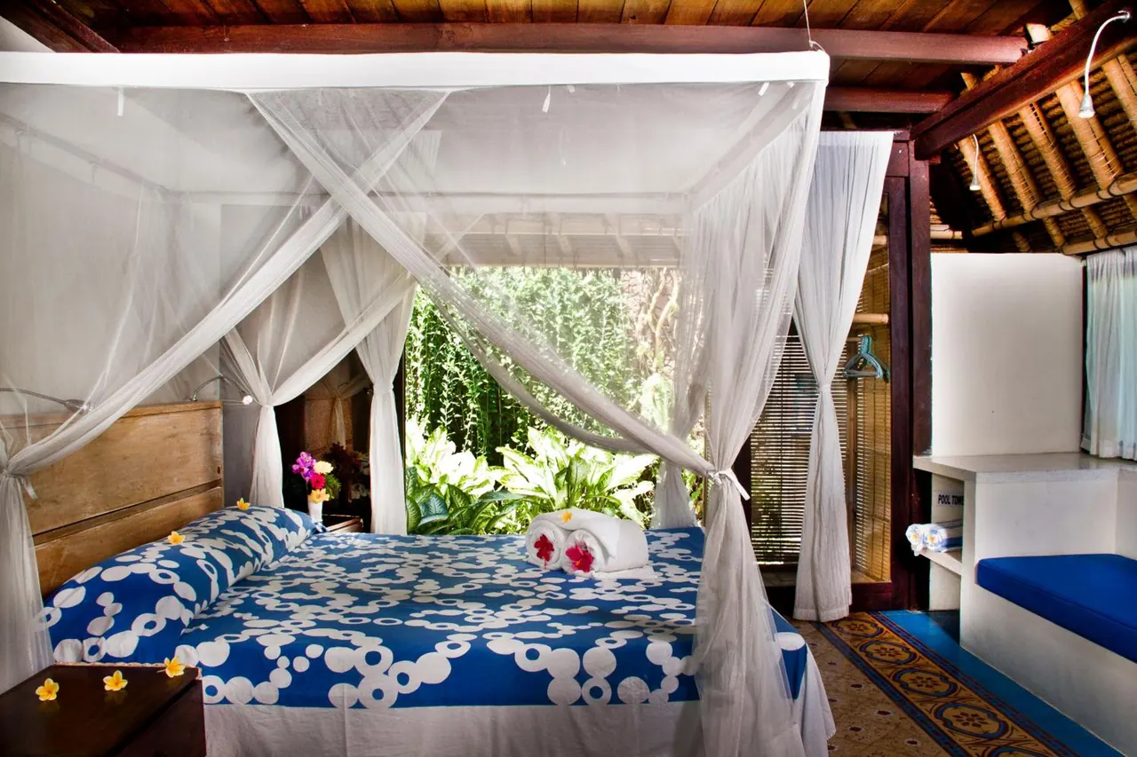 A canopied double bed with blue and white bedding, folded towels and flowers, beside a window opening onto tropical garden greenery.