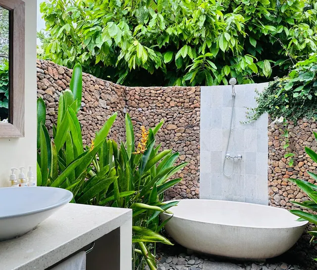An outdoor bathroom with a freestanding oval bathtub, an open tiled shower and a vessel sink beside a stone wall and tropical plants.