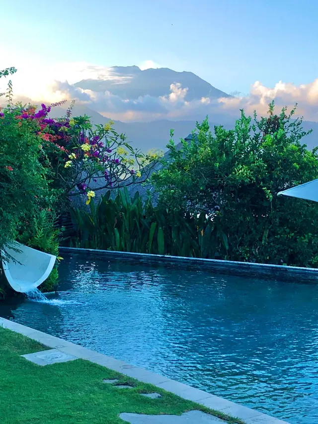 A swimming pool with a curved white water spout beside it, flowering shrubs and greenery, and a cloud-covered mountain in the background.
