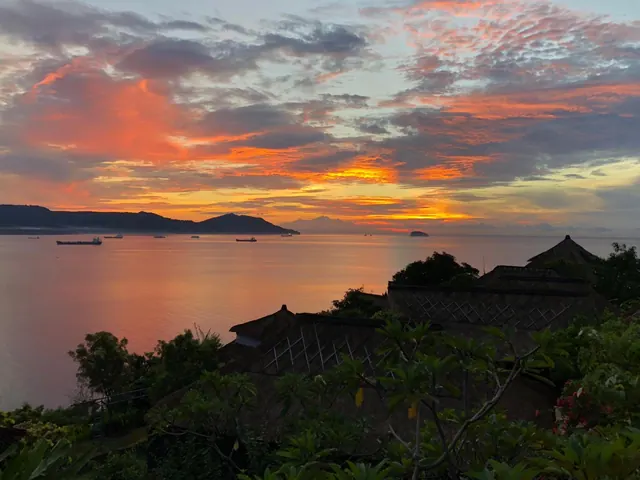 A colourful sky over a calm sea with anchored cargo ships, distant mountains and thatched roofs in the foreground.