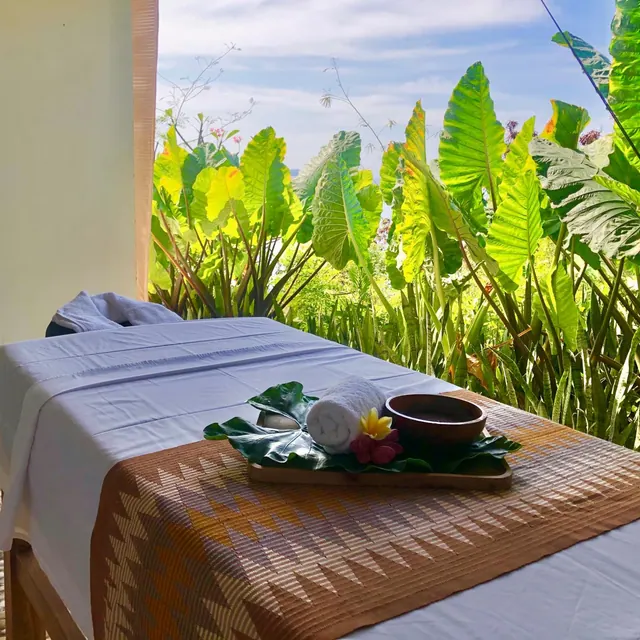 A massage table with a folded towel and a leaf-lined tray holding flowers and a bowl, with tropical garden plants and sky visible beyond the open side of the room.