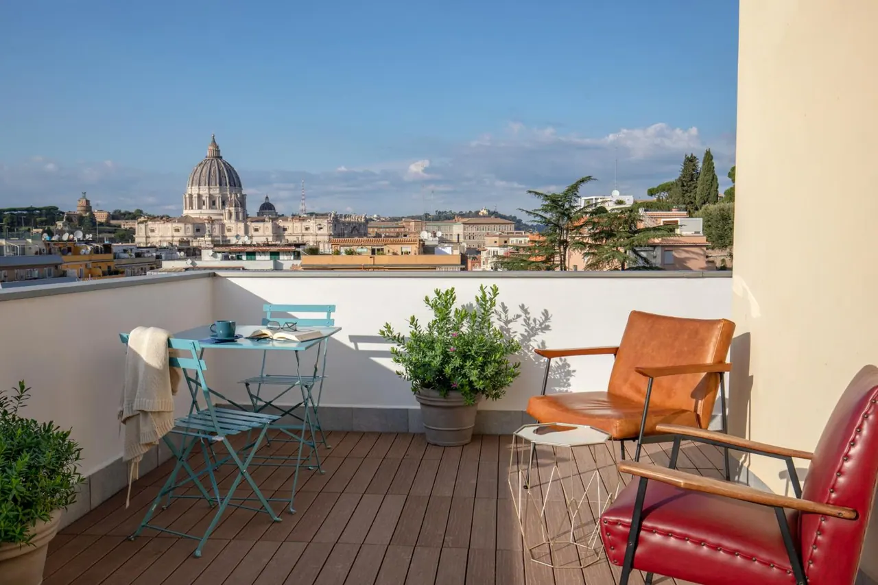 A rooftop terrace with a bistro table, chairs and potted plants, overlooking the city with the dome of St. Peter's Basilica in the distance.