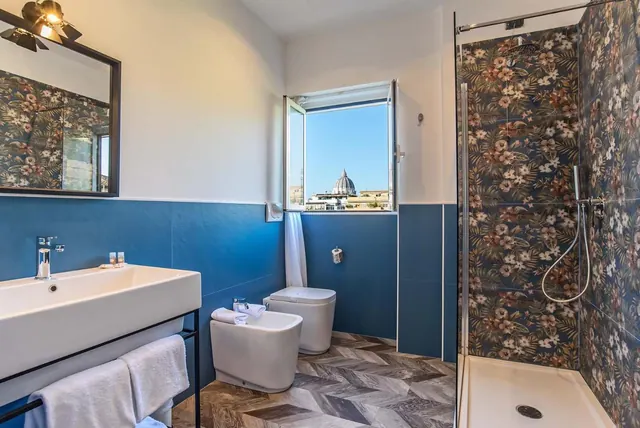 A bathroom with a sink on a black metal frame, a bidet and toilet, a walk-in shower with floral tiles, and a window showing the dome of St. Peter's Basilica over rooftops.