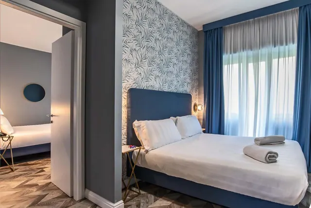 A bedroom with a made double bed, blue headboard and curtains, patterned wallpaper, and an open door showing a second room.