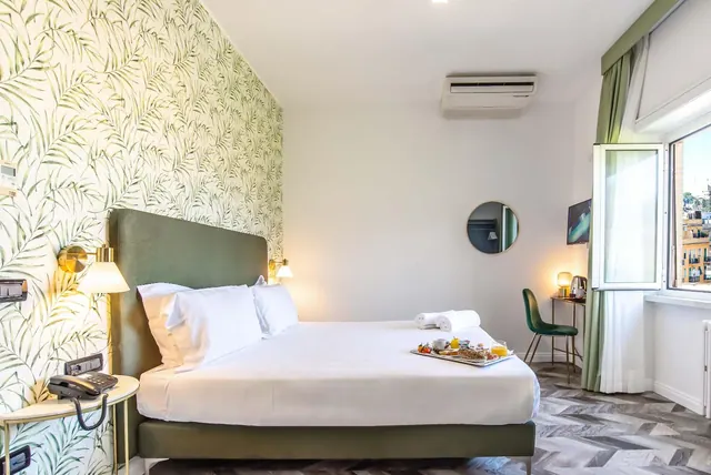 A bedroom with green leaf-patterned wallpaper, a double bed with a breakfast tray, a wall-mounted air conditioner, a round mirror and a window looking onto neighbouring buildings.
