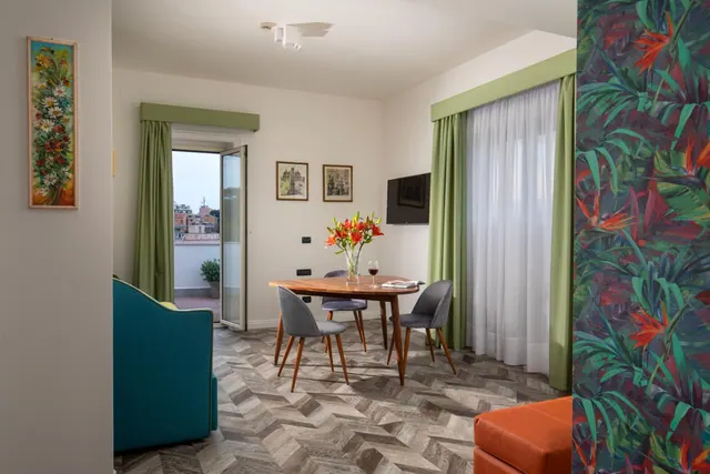 A staged living and dining area with a round wooden table, grey chairs, a teal armchair, and an open balcony door showing rooftops of the city.