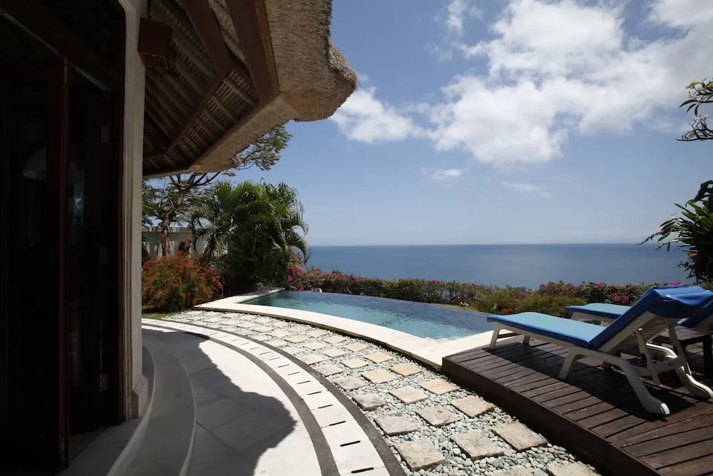 A small pool beside a thatched-roof villa with blue sun loungers on a timber deck, overlooking the sea.