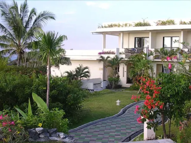A white two-storey building with upper balconies stands behind a lawn, a checkered paved path and tropical plants including palm trees and flowering shrubs.