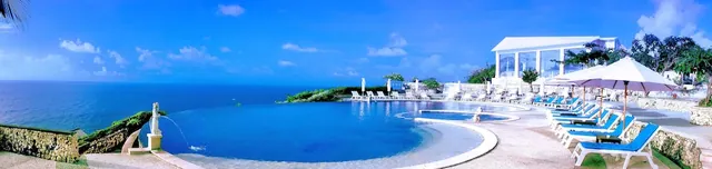 An infinity-edge pool with white sun loungers and parasols overlooks the ocean, with a white pavilion and a statue fountain beside it.