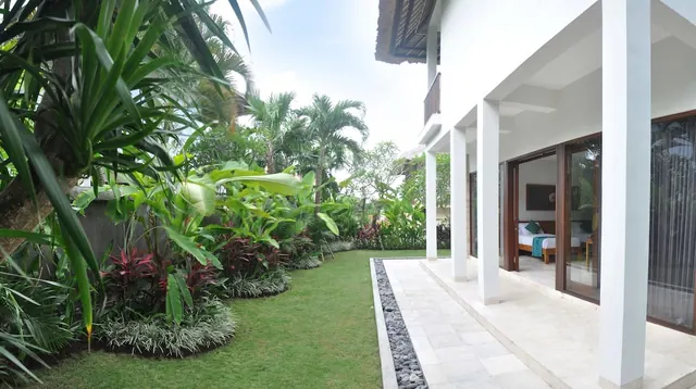 A landscaped lawn with tropical plants and palm trees runs beside a white villa with a tiled terrace, and an open door shows a bedroom with a double bed.