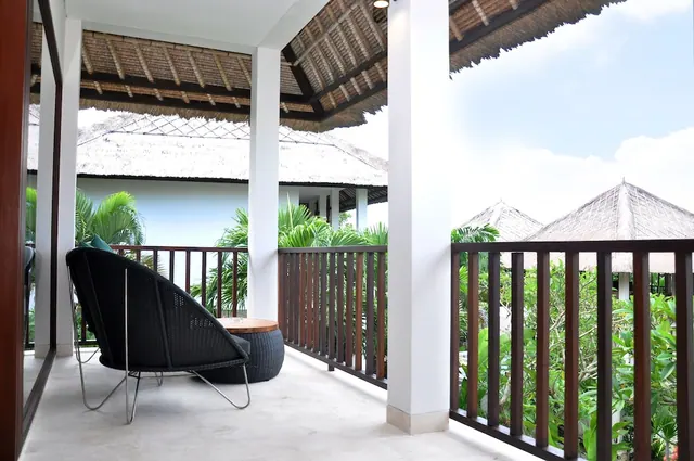 A covered balcony with a black wicker lounge chair and a wooden side table, with wooden railings overlooking greenery and thatched roofs.