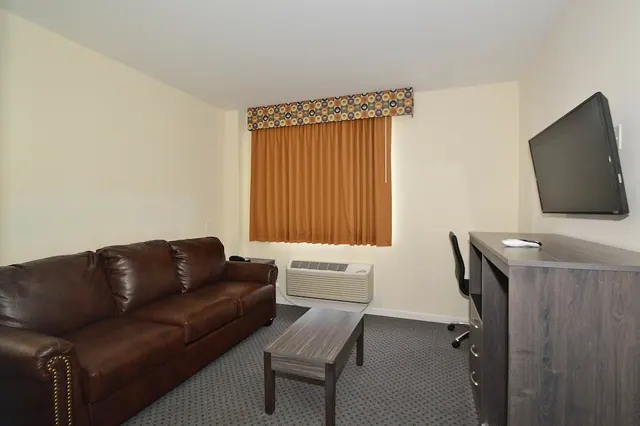 A living area with a brown leather sofa, a wooden coffee table, a wall-mounted flat-screen TV, a wooden dresser, and a window with orange curtains and a wall air conditioner beneath it.