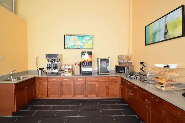 A hotel breakfast area with a coffee machine, juice dispenser, mini fridge, microwave and pastry display on a countertop along yellow walls.