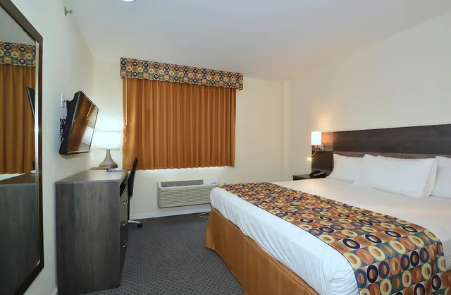 A made king bed sits beside a wall-mounted television, a desk with a lamp and chair, and a wall air conditioning unit under a curtained window.