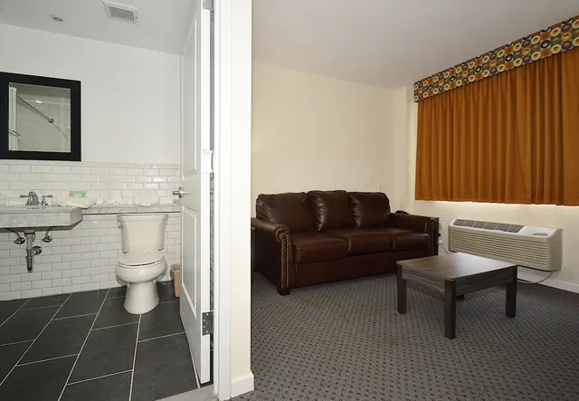 A living area with a brown leather sofa, a wooden coffee table, orange curtains and a wall air conditioning unit, with an open bathroom door showing a toilet and sink on the left.