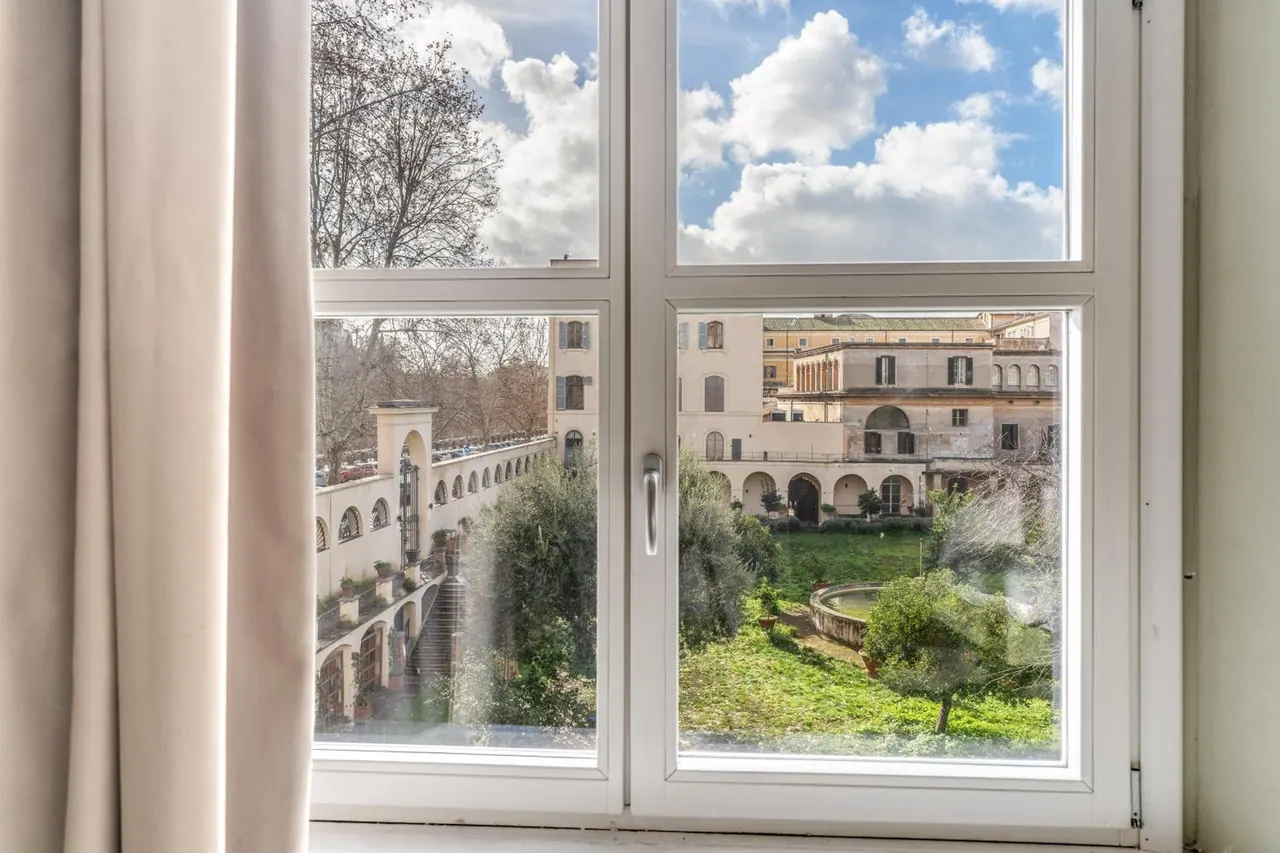 A white-framed window with beige curtains looks out onto a green garden courtyard surrounded by historic arched buildings.