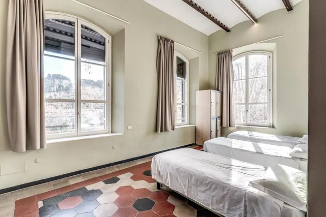 A bedroom with single beds, tall arched windows looking out onto trees, a tan wardrobe and a hexagonal tiled floor.