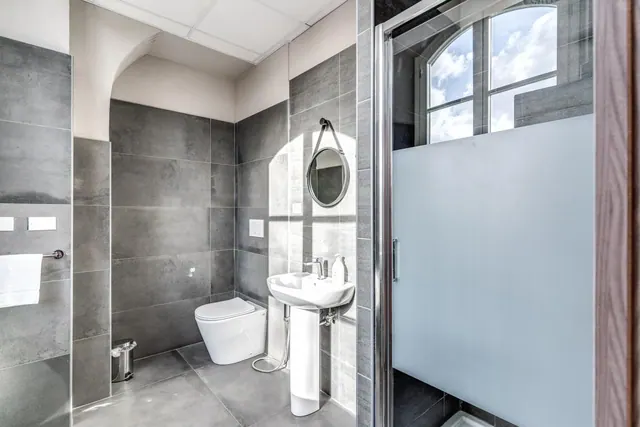 A bathroom with grey tiled walls, a wall-hung toilet, a pedestal sink, a round mirror and a frosted glass walk-in shower, with an arched window above the shower.
