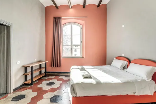 A bedroom with a double bed with white linen, a terracotta-coloured wall with an arched window, and a hexagonal tiled floor.