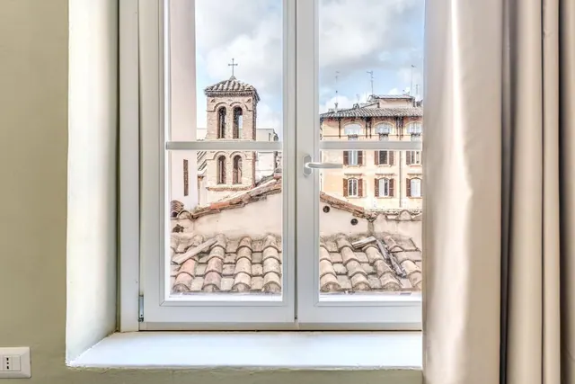 A white double-casement window opens onto terracotta rooftops, a brick bell tower topped with a cross, and cream-coloured apartment buildings under a cloudy sky.