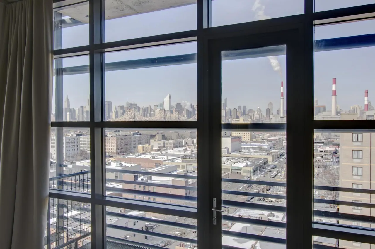 View through floor-to-ceiling windows and a glass door over a city skyline, with a bridge, smokestacks and low-rise buildings below, and a curtain on the left.