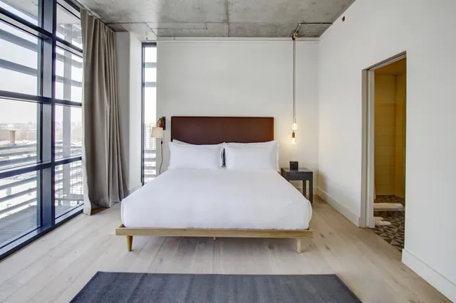 A bedroom with a made king bed, floor-to-ceiling windows showing buildings outside, and an open doorway to a bathroom.
