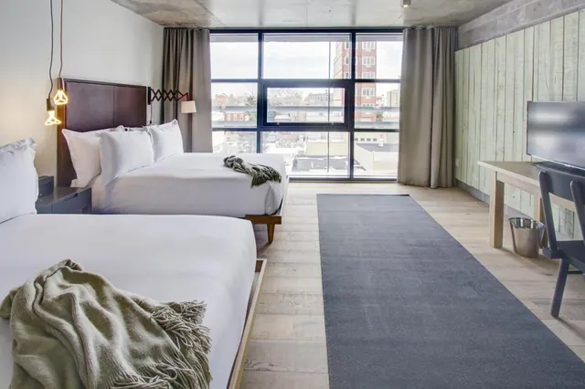 A hotel room with two beds with white linens, a dark grey rug, a wooden desk with a television, and large windows looking out onto neighbouring buildings.