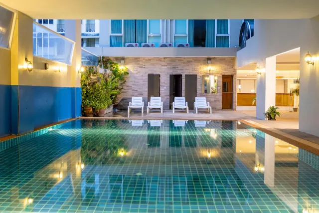 An indoor swimming pool at dusk with four white sun loungers along the far edge and potted plants beside the wall.