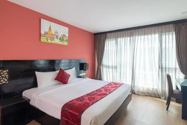 A bedroom with a king bed, red bed runner and red accent wall, with a work desk beside floor-to-ceiling windows covered in sheer curtains.