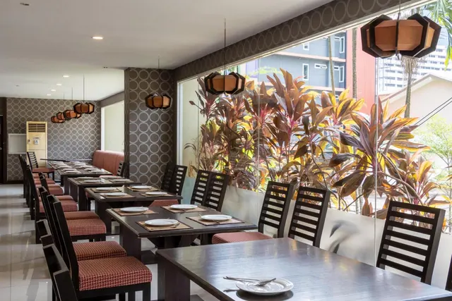 A restaurant dining room with set tables, dark wooden chairs with red cushions, pendant lamps, and large windows looking onto tropical plants and buildings outside.