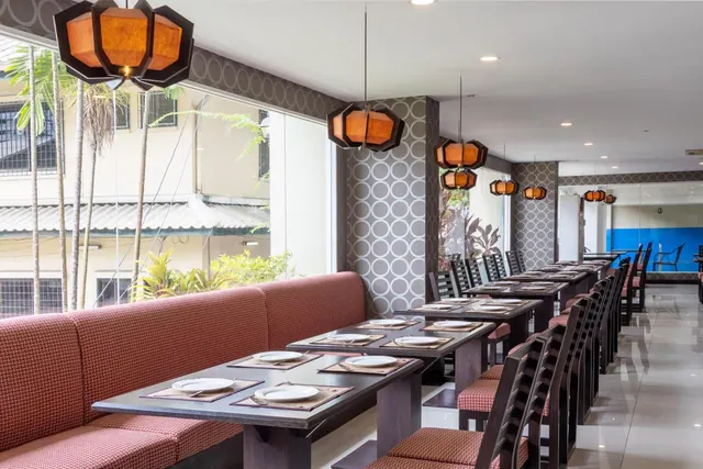 A restaurant dining room with long tables set with plates, a red upholstered banquette along the windows, and pendant lanterns hanging overhead.