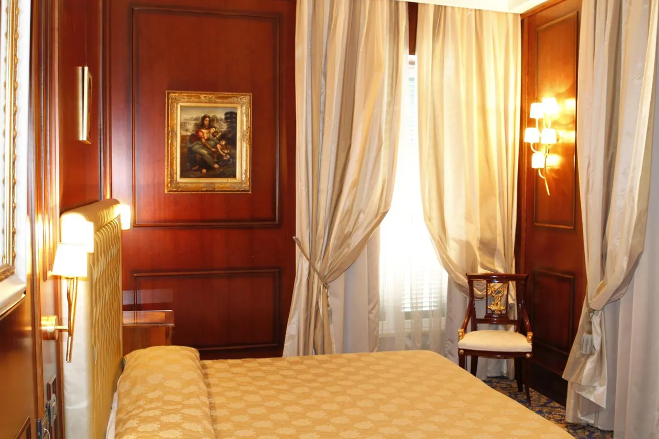 A bedroom with a double bed under a gold bedspread, wood-panelled walls, silk curtains, a framed painting and a wooden chair by the window.