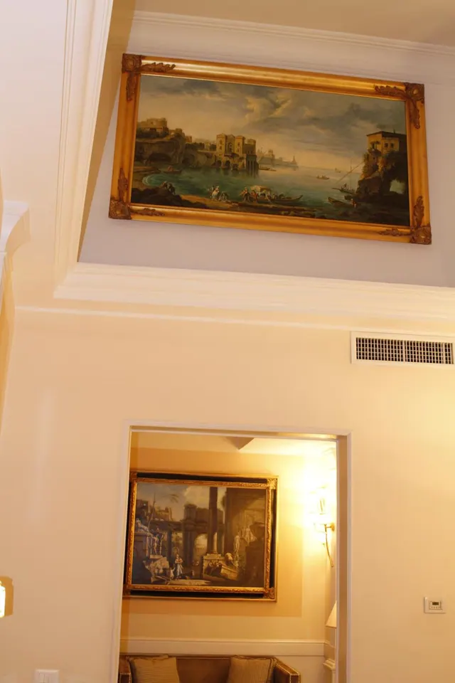 A large framed painting of a coastal scene hangs on a cream wall above a doorway, with a second framed painting and a sofa visible in the room beyond.